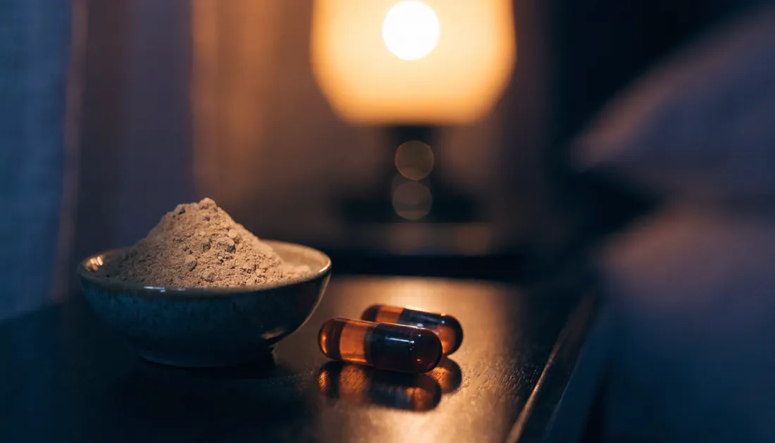 Ashwagandha root powder in a bowl with capsules on a dark nightstand
