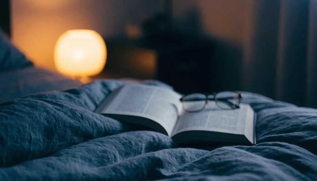 An open book and reading glasses on a bed beside a warm bedside lamp