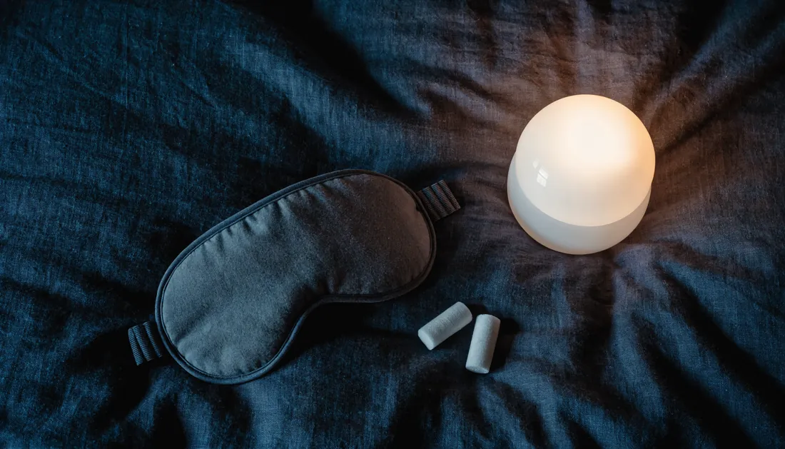 A sleep eye mask, earplugs, and a small bedside light on dark linen